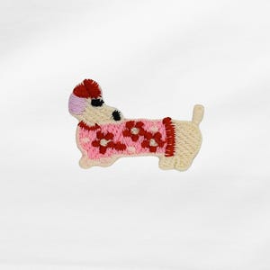 Könnte beinhalten: Gestickter Aufnäher eines Dackelhundes mit einem rosa Pullover mit roten Blumenakzenten. Der Hund hat einen roten und lavendelfarbenen Hut, schwarze Augen und einen beigefarbenen Körper. Der Aufnäher ist ein niedliches, skurriles Design.