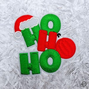 Könnte beinhalten: Gestickter Aufnäher mit den Worten "HO HO" in Grün und Rot. Das Design beinhaltet einen Weihnachtsmannhut und eine rote Kugel. Der Aufnäher befindet sich auf weißem Hintergrund.