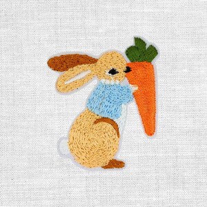 Könnte beinhalten: Gestickter Aufnäher mit einem Hasen in einer blauen Jacke, der eine große orangefarbene Karotte mit grünen Blättern hält. Der Hase hat hellbraunes Fell, braune Ohren und schwarze Augen. Auf weißem Stoffhintergrund.