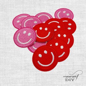 Könnte beinhalten: Eine Ansammlung runder, bestickter Aufnäher mit Smiley-Gesichtern in Rot- und Rosatönen. Jeder Aufnäher hat einen weißen gestickten Rand, zwei weiße runde Augen und einen weißen geschwungenen Mund, angeordnet auf einem strukturierten Stoff.