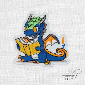 Könnte beinhalten: Gestickter Aufnäher mit einem blauen Drachen, der ein gelbes Buch liest. Der Drache hat orange Augen, grüne Kopfschuppen und braune und orangefarbene Flügel. Der Aufnäher hat einen weißen Rand und den Text "material4 DIY".