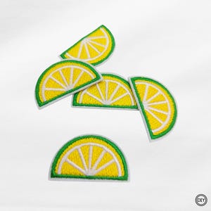 Könnte beinhalten: Gestickte Aufnäher in Form von gelb-weißen Zitronenscheiben mit grünen Rändern. Die Aufnäher sind auf weißem Hintergrund angeordnet. DIY-Bastelartikel.