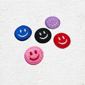 Könnte beinhalten: Fünf runde, gestickte Aufnäher mit Smiley-Gesichtern. Die Aufnäher sind in den Farben Blau, Schwarz, Lila, Rot und Rosa gehalten, jeweils mit einem weißen, aufgestickten Lächeln.