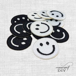 Könnte beinhalten: Eine Sammlung runder, bestickter Aufnäher mit Smiley-Gesichtern in Schwarz und Weiß. Die Aufnäher sind in einer Gruppe angeordnet und zeigen abwechselnd schwarze Gesichter mit weißer Stickerei und weiße Gesichter mit schwarzer Stickerei.