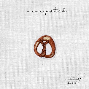 Könnte beinhalten: Ein kleiner gestickter Brezel-Aufnäher mit braunen und beigen Garnfarben. Das Design zeigt eine traditionelle verschlungene Brezelform mit einem weißen Rand. Der Text mini patch steht darüber und material4 DIY steht darunter.