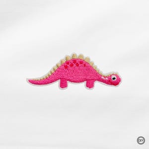 Könnte beinhalten: Gestickter Aufnäher mit einem rosa Dinosaurier mit cremefarbenem Rücken und Akzenten. Der Dinosaurier hat einen weißen Rand und ein kleines Auge. Geeignet für Kleidung, Taschen oder andere Stoffartikel.