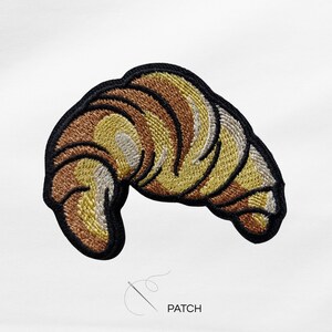 Könnte beinhalten: Gestickter Aufnäher in Form eines Croissants, mit Braun-, Gelb- und Cremetönen, umrandet von einem schwarzen Rand. Das Wort "PATCH" ist unter dem Bild aufgedruckt, zusammen mit einer Nadel- und Fadenillustration.
