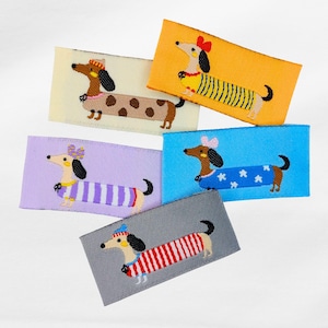 Könnte beinhalten: Fünf rechteckige Stoffaufnäher mit gestickten Dackel-Designs. Die Hunde sind in verschiedenen Outfits gekleidet, wie gestreiften Pullovern und gepunkteten Mänteln, vor Hintergründen in Gelb, Blau, Lavendel, Grau und Creme.