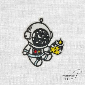 Könnte beinhalten: Gestickter Aufnäher mit einem Cartoon-Astronauten in einem weißen Raumanzug mit einem schwarzen Helm, der eine sternenklare Nachtszene zeigt. Der Astronaut hält einen Beutel mit gelben Sternen. Der Aufnäher hat eine schwarze Umrandung und rote und blaue Akzente.