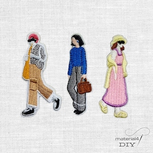 Könnte beinhalten: Drei gestickte Aufnäher mit stilisierten Figuren. Die erste trägt eine rote Mütze, ein kariertes Hemd und eine beige Hose. Die zweite hat dunkle Haare, einen blauen Pullover und eine graue Hose mit einer braunen Tasche. Die dritte trägt einen gelben Hut und ein rosa Kleid.