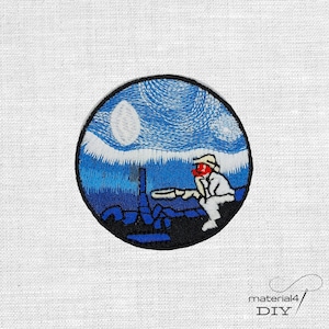Könnte beinhalten: Gestickter Aufnäher mit einem kreisförmigen Design, das einen Sternenhimmel in Blau und Weiß darstellt. Eine Figur in weißer Kleidung und mit roter Gesichtsbedeckung sitzt und betrachtet eine Struktur. Der Aufnäher hat einen schwarzen Rand.