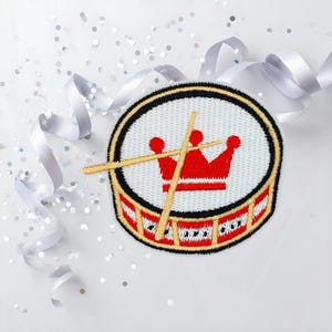 Könnte beinhalten: Gestickter Aufnäher mit einem Trommeldesign mit roter Krone und Trommelstöcken. Der Aufnäher hat einen weißen Hintergrund mit schwarzem und goldenem Rand. Weißes Band und silbernes Konfetti sind um den Aufnäher verstreut.
