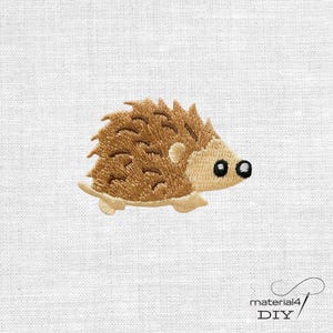 Könnte beinhalten: Ein gestickter Aufnäher, der einen braunen Igel mit hellbeigem Gesicht, kleinen abgerundeten Ohren sowie einer schwarzen Nase und einem schwarzen Auge zeigt. Das Motiv ist auf einen weißen Stoff gestickt. Der Text material4 DIY ist unten rechts sichtbar.