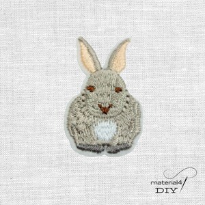 Könnte beinhalten: Ein gestickter Aufnäher, der einen grauen Hasen mit langen Ohren, braunen Augen, einer kleinen braunen Nase und einem weißen Bauchfleck zeigt. Der Hase ist in sitzender Position dargestellt, mit detaillierten Stichen im Fell und Gesicht.