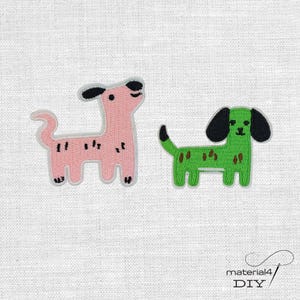Könnte beinhalten: Zwei gestickte Hundeaufnäher. Einer ist rosa mit schwarzen Flecken und Ohren, der andere ist grün mit schwarzen Ohren und braunen Flecken. Beide haben weiße Umrisse und befinden sich auf einem weißen Stoffhintergrund. Die Aufnäher sind für DIY-Projekte.