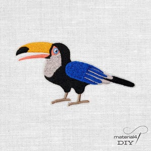 Könnte beinhalten: Gestickter Toucan-Aufnäher mit gelbem Schnabel, blauen Flügeln und schwarzem Körper. Der Vogel steht auf zwei braunen Beinen vor einem weißen Hintergrund. Der Aufnäher ist ideal für DIY-Projekte oder zur Dekoration von Kleidung.