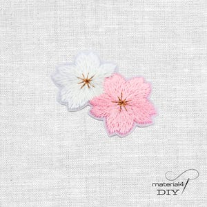 Könnte beinhalten: Zwei gestickte Blumenaufnäher, einer weiß und einer rosa, mit braunen Zentren. Die Aufnäher befinden sich auf einem weißen Stoffhintergrund. Der Text "material4 DIY" befindet sich in der unteren rechten Ecke.