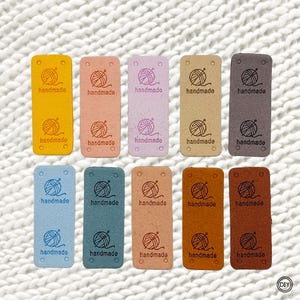 Könnte beinhalten: Zehn rechteckige Etiketten in verschiedenen Farben, darunter Gelb, Rosa und Braun. Jedes Etikett trägt das Wort "handmade" und eine Grafik von Garn und Häkelhaken. Die Etiketten haben kleine Löcher zum Befestigen an Artikeln.
