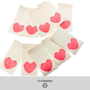 Könnte beinhalten: Eine Sammlung von zehn cremefarbenen Stoffetiketten, jedes mit einem leuchtend roten Herzmotiv. Die Etiketten sind auf weißem Hintergrund angeordnet. Der Text "10 Etiketten" ist unten sichtbar.