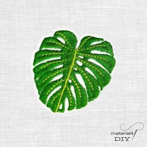 Könnte beinhalten: Ein gestickter Aufnäher in Form eines Monstera-Blattes mit leuchtend grünem Garn und detaillierten gelben Nähten entlang der Blattadern. Das Design weist eine ausgeprägte Blattstruktur mit charakteristischen Ausschnitten auf.