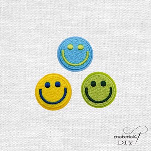 Könnte beinhalten: Drei kreisförmige gestickte Aufnäher mit Smiley-Gesichtern. Ein Aufnäher ist hellblau mit einem lindgrünen Gesicht, einer ist gelb mit einem dunkelblauen Gesicht und einer ist lindgrün mit einem dunkelgrünen Gesicht, alle mit gestickten Rändern.