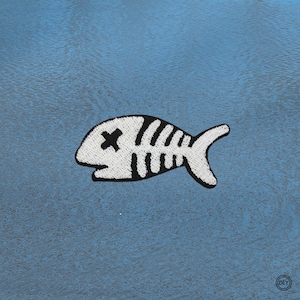 Könnte beinhalten: Gestickter Aufnäher mit einem stilisierten Fischskelett-Design. Der Aufnäher ist weiß mit schwarzen Details, einschließlich eines 'X' für ein Auge. Der Hintergrund ist eine strukturierte, hellblaue Oberfläche. Für DIY-Projekte.
