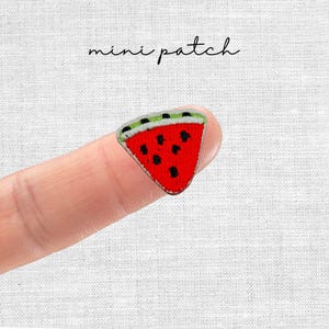 Könnte beinhalten: Ein kleiner, gestickter Wassermelonen-Aufnäher. Der Aufnäher ist ein rotes Dreieck mit schwarzen Samen und einer grünen und weißen Schale. Die Worte "mini patch" sind in Kursivschrift darüber geschrieben.