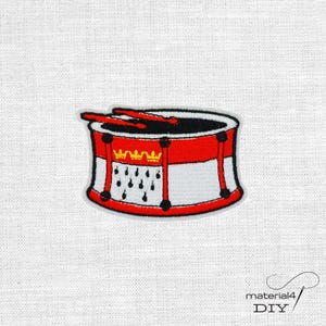 Könnte beinhalten: Ein gestickter Aufnäher, der eine rot-weiße Trommel mit zwei darauf liegenden roten Trommelstöcken zeigt. Das Trommeldesign enthält eine gelbe Krone und schwarze Hermelinmuster auf dem weißen Bereich, umrandet von einem dunklen Rand.