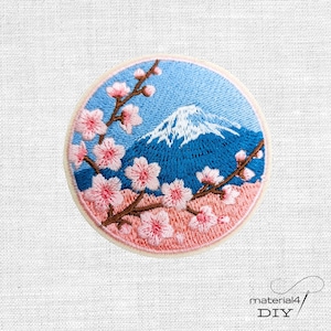 Könnte beinhalten: Ein runder Stickaufnäher mit einem blauen Berg mit weißer Schneekuppe, umrahmt von braunen Zweigen mit hellrosa Kirschblüten. Das Motiv ist vor einem hellblauen Himmelshintergrund mit einer zartrosa Basis dargestellt.