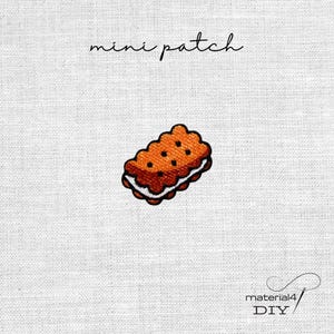 Könnte beinhalten: Ein gestickter Mini-Aufnäher in Form eines rechteckigen braunen Sandwich-Kekses mit weißer Cremefüllung und kleinen dunklen Punkten. Der Text mini patch steht über dem Design und material4 DIY steht darunter.
