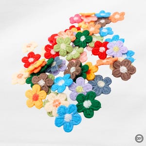 Könnte beinhalten: Eine Sammlung kleiner, gestickter Blumenapplikationen in verschiedenen Farben, darunter Rot, Blau, Grün, Gelb und Lila. Jede Blume hat eine kontrastierende Mitte, wodurch ein lebendiges und strukturiertes Aussehen entsteht. Diese DIY-Bastelmaterialien sind ideal zum Verzieren von Kleidung oder Accessoires.