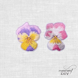 Könnte beinhalten: Zwei gestickte Stiefmütterchen-Blumenaufnäher. Einer hat lila und gelbe Blütenblätter, der andere rosa und weiße. Die Aufnäher befinden sich auf einem weißen Stoffhintergrund. Ideal für DIY-Projekte.