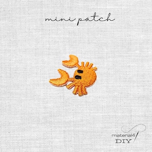 Könnte beinhalten: Ein orangefarbener, gestickter Krabbenaufnäher mit schwarzen Augen und zwei großen Scheren. Der Text mini patch steht über der Krabbe und der Text material4 DIY befindet sich in der unteren rechten Ecke auf einem weißen, strukturierten Stoff.