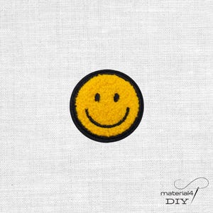 Könnte beinhalten: Ein kreisförmiger gestickter Aufnäher mit einem leuchtend gelben Smiley-Gesicht, einer schwarzen gestickten Umrandung sowie schwarzen Augen und einem geschwungenen Mund. Die Textur des gelben Garns ist flauschig und erhaben, was einen Kontrast zum schwarzen Rand bildet.