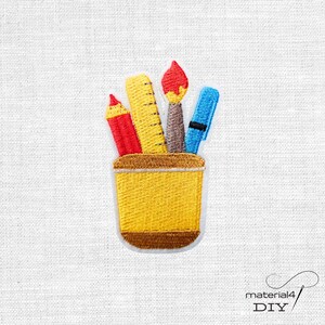 Könnte beinhalten: Gestickter Aufnäher mit einem gelben Stifthalter, gefüllt mit Schulmaterial. Der Aufnäher zeigt einen roten Bleistift, ein gelbes Lineal, einen braunen Pinsel und einen blauen Stift, vor weißem Hintergrund.