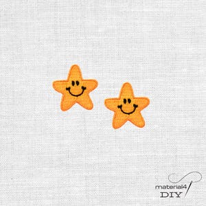 Könnte beinhalten: Zwei orangefarbene, sternförmige Stickaufnäher mit einem aufgestickten schwarzen Smiley-Gesicht. Jeder Stern hat fünf Zacken und einen fröhlichen Ausdruck mit zwei kleinen schwarzen Augen und einem geschwungenen schwarzen Mund.