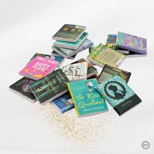 Könnte beinhalten: Eine Sammlung von Miniaturbüchern mit verschiedenen bunten Einbänden, darunter Titel wie Happy Place, The Assassin's Blade, The Kiss Quotient und Little Women, die auf einer Oberfläche verstreut sind, umgeben von kleinen goldenen Glitzerpartikeln.