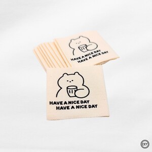 Könnte beinhalten: Ein Stapel cremefarbener Stoffetiketten. Jedes Etikett zeigt eine schwarze Strichzeichnung eines Bären, der eine Tasse hält. Der Text "HAVE A NICE DAY" ist in Schwarz unter der Bärenillustration gedruckt.