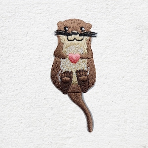 Könnte beinhalten: Gestickter Aufnäher eines Cartoon-Otters, der ein rosa Herz hält. Der Otter hat braunes Fell, einen cremefarbenen Bauch und schwarze Schnurrhaare. Der Aufnäher ist zum Aufbügeln oder Aufnähen auf Stoff gedacht.