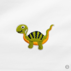 Könnte beinhalten: Ein gestickter Aufbügler in Form eines Cartoon-Dinosauriers mit hellgrünem Körper, dunkelblauen vertikalen Streifen auf dem Rücken und einem orangefarbenen Bauch. Der Dinosaurier hat ein weißes Auge mit schwarzer Pupille und ein kleines schwarzes Lächeln.