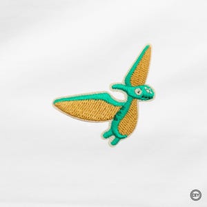 Könnte beinhalten: Gestickter Aufnäher mit einem fliegenden Pterodactylus. Der Aufnäher hat einen beigefarbenen Körper mit türkisfarbenen Konturen und Akzenten. Ideal für DIY-Projekte und Bastelarbeiten.