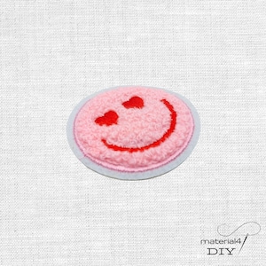Könnte beinhalten: Ein kreisförmiger gestickter Aufnäher mit einer strukturierten, hellrosa, flauschigen Oberfläche. Der Aufnäher zeigt ein rotes, gesticktes Smiley-Gesicht mit zwei kleinen roten herzförmigen Augen und einem geschwungenen roten Mund, weiß umrandet.