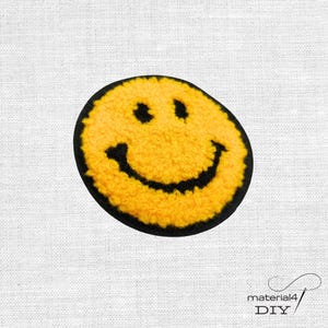Könnte beinhalten: Ein kreisförmiger Aufnäher mit einem gelben, flauschigen Smiley-Gesicht, schwarzen gestickten Augen und einem breiten, geschwungenen schwarzen Mund. Das Design ist von einem soliden schwarzen Rand umgeben, der einen kontrastreichen Akzent setzt.
