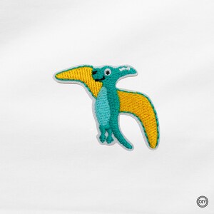 Könnte beinhalten: Gestickter Aufnäher eines türkis-gelben Pterodactylus. Der Pterodactylus hat einen türkisfarbenen Körper, gelbe Flügel und ein kleines, lächelndes Gesicht. Der Aufnäher ist weiß umrandet und wahrscheinlich für Kleidung oder Accessoires.