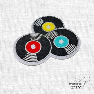 Könnte beinhalten: Gestickte Aufnäher mit drei Schallplatten mit schwarzen Hintergründen und farbigen Zentren in Rot, Gelb und Türkis. Jede Platte hat silberfarbene Rillen und einen weißen Rand. Die Aufnäher sind in einer Gruppe angeordnet.