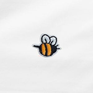 Könnte beinhalten: Gestickter Aufnäher einer Cartoon-Biene. Die Biene hat einen gelb-schwarz gestreiften Körper, schwarze Flügel und einen schwarzen Kopf. Der Aufnäher ist grau umrandet und befindet sich auf weißem Hintergrund.