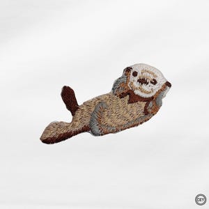 Könnte beinhalten: Gestickter Aufnäher mit einem Seeotter in Braun-, Beige- und Grautönen. Der Otter liegt auf dem Rücken, die Pfoten am Gesicht. Der Aufnäher hat ein detailliertes, strukturiertes Aussehen, ideal für Kleidung oder Accessoires.