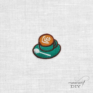 Könnte beinhalten: Ein gestickter Aufnäher mit einer blaugrünen Kaffeetasse, braunem Latte-Art-Muster und einem weißen Löffel auf der Untertasse. Das Motiv ist mit dunkelbraunen Umrandungen auf strukturiertem weißem Stoff gestickt, inklusive dem Text material4 DIY.