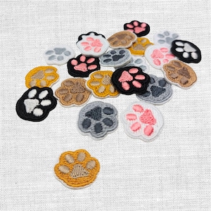 Könnte beinhalten: Eine Sammlung gestickter Pfotenabdruck-Patches in verschiedenen Farben, darunter Schwarz, Weiß, Rosa, Braun, Grau und Gelb. Jeder Patch zeigt ein detailliertes Pfotenballen-Design mit weißem Rand. Diese Patches eignen sich zum Verzieren von Kleidung oder Accessoires.