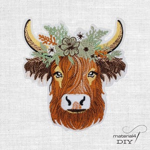 Könnte beinhalten: Ein gestickter Aufnäher mit einem Highland-Rinderkopf mit langen, geschwungenen Hörnern, braunem Fell und einem Kranz aus grünen Blättern und Blumen in Weiß-, Orange- und Brauntönen. Das Design zeigt detaillierte Stickereien und einen weißen Rand.